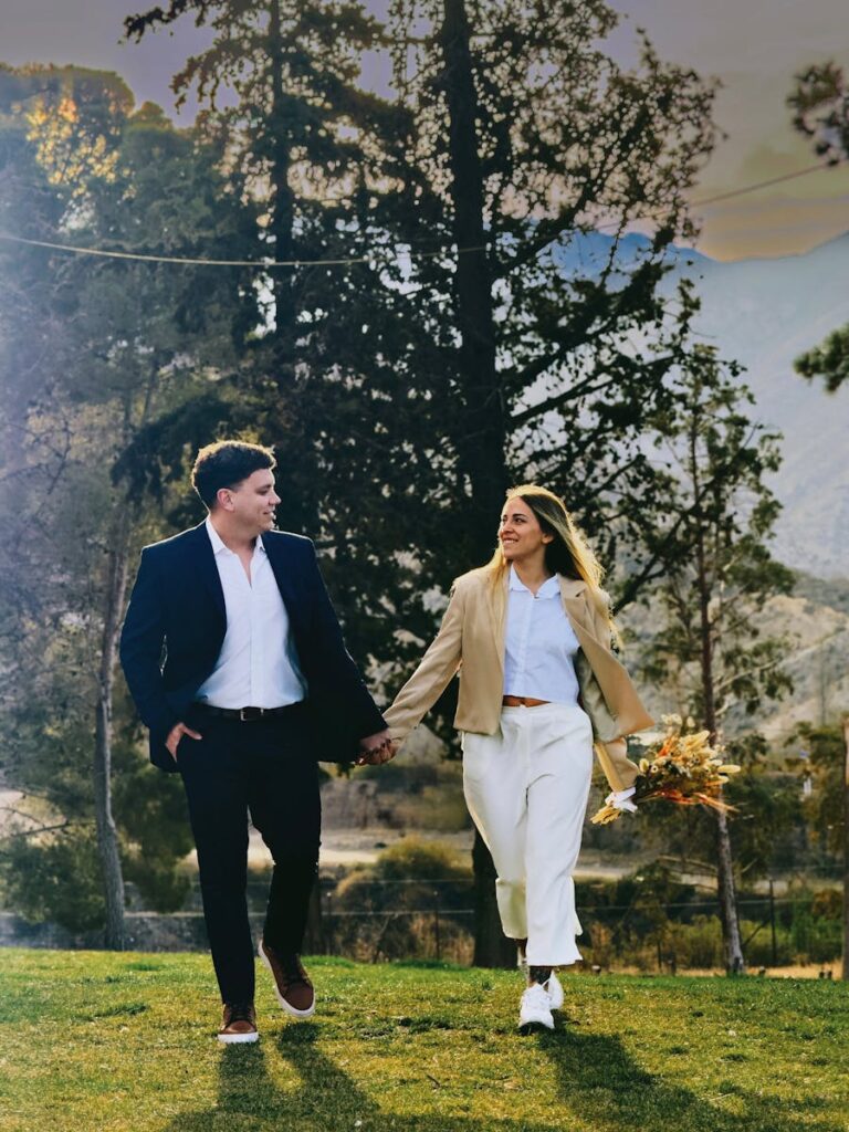 A couple holding hands wearing a womens trouser suit & mens suit and walking together in a beautiful park setting during fall.