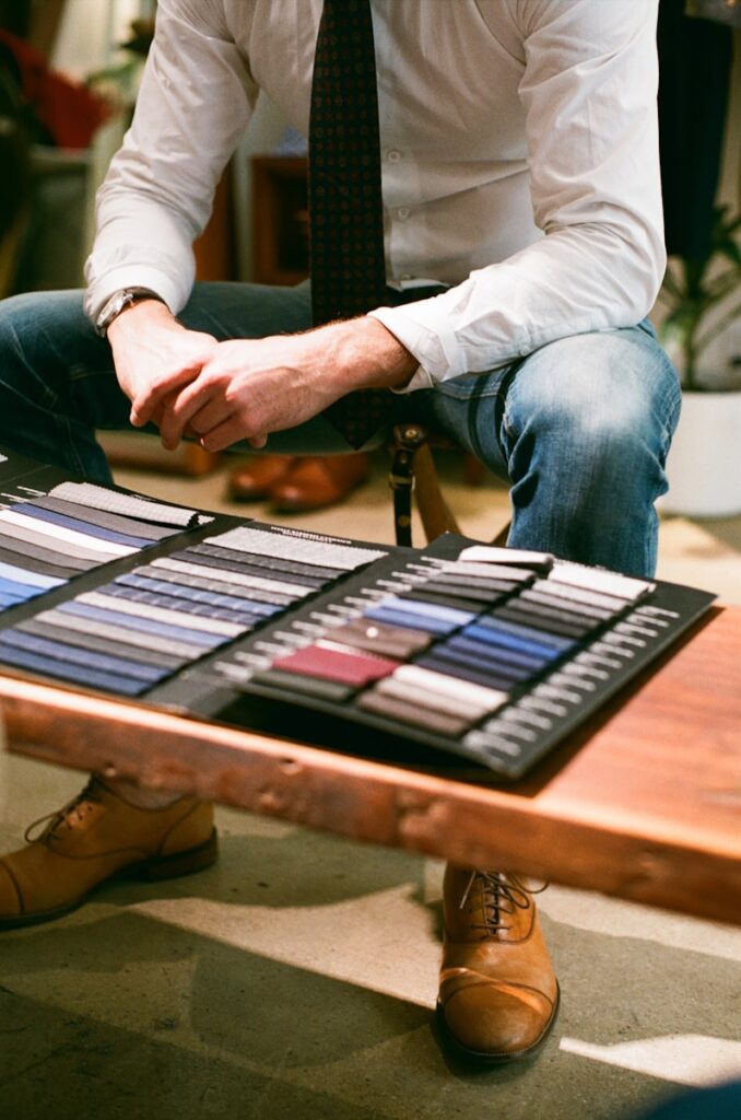 a man sitting at a table with a display of ties on it contemplating bespoke suit vs made to measure